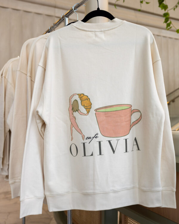 White sweatshirt on a hanger featuring a pink coffee cup illustration and the text OLIVIA beneath it.