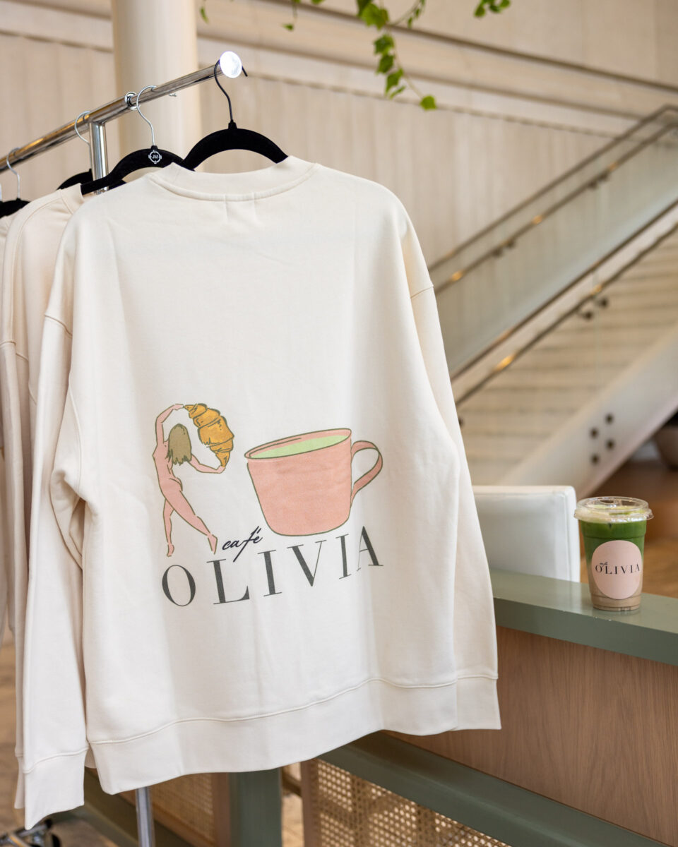 White sweatshirt on a hanger with a pink cup illustration and the word OLIVIA, in a store display background with drinks nearby